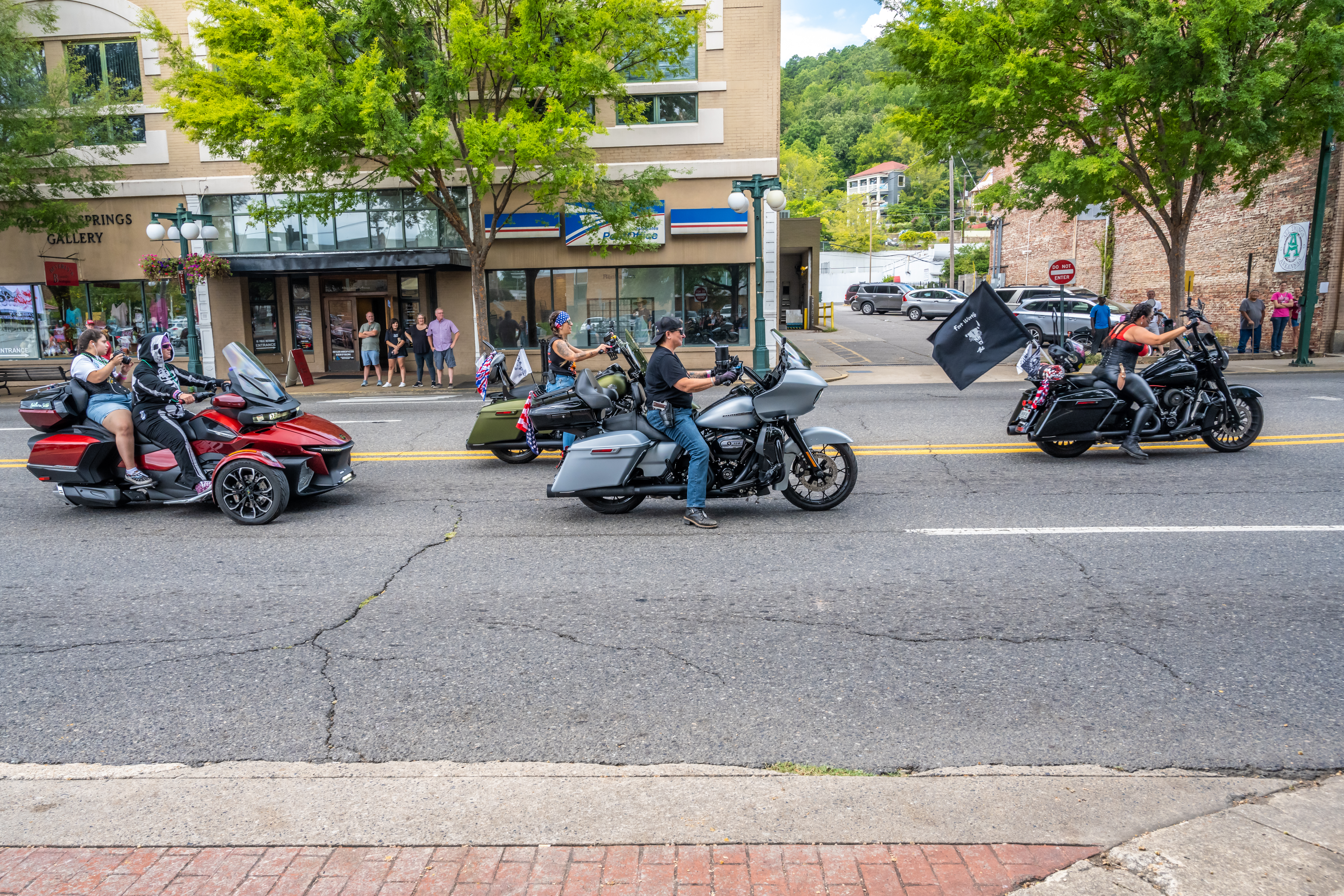 Motorcyclists Passing Local Shops in Hot Springs A line of motorcyclists, including a rider on a red Can-Am Spyder and others on touring bikes, riding past a U.S. Post Office and historic storefronts in Hot Springs.