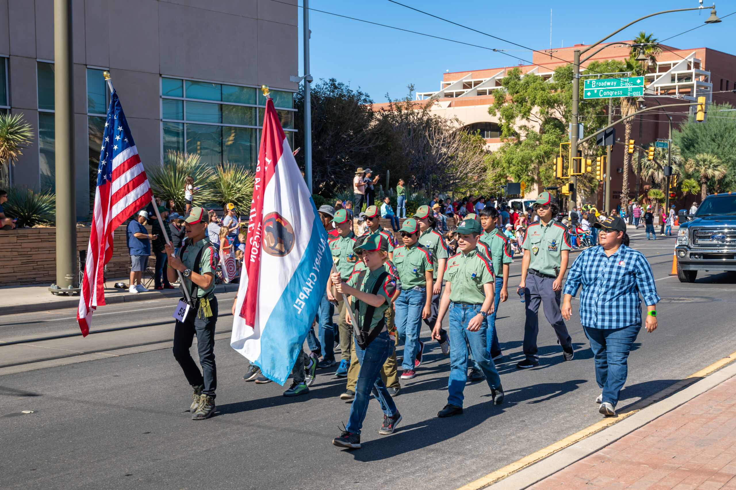Calvary Chapel Tucson Youth Parade March A youth group in olive green scout-style shirts marching in a Tucson parade, carrying an American flag and a blue and white Calvary Chapel Tucson flag.