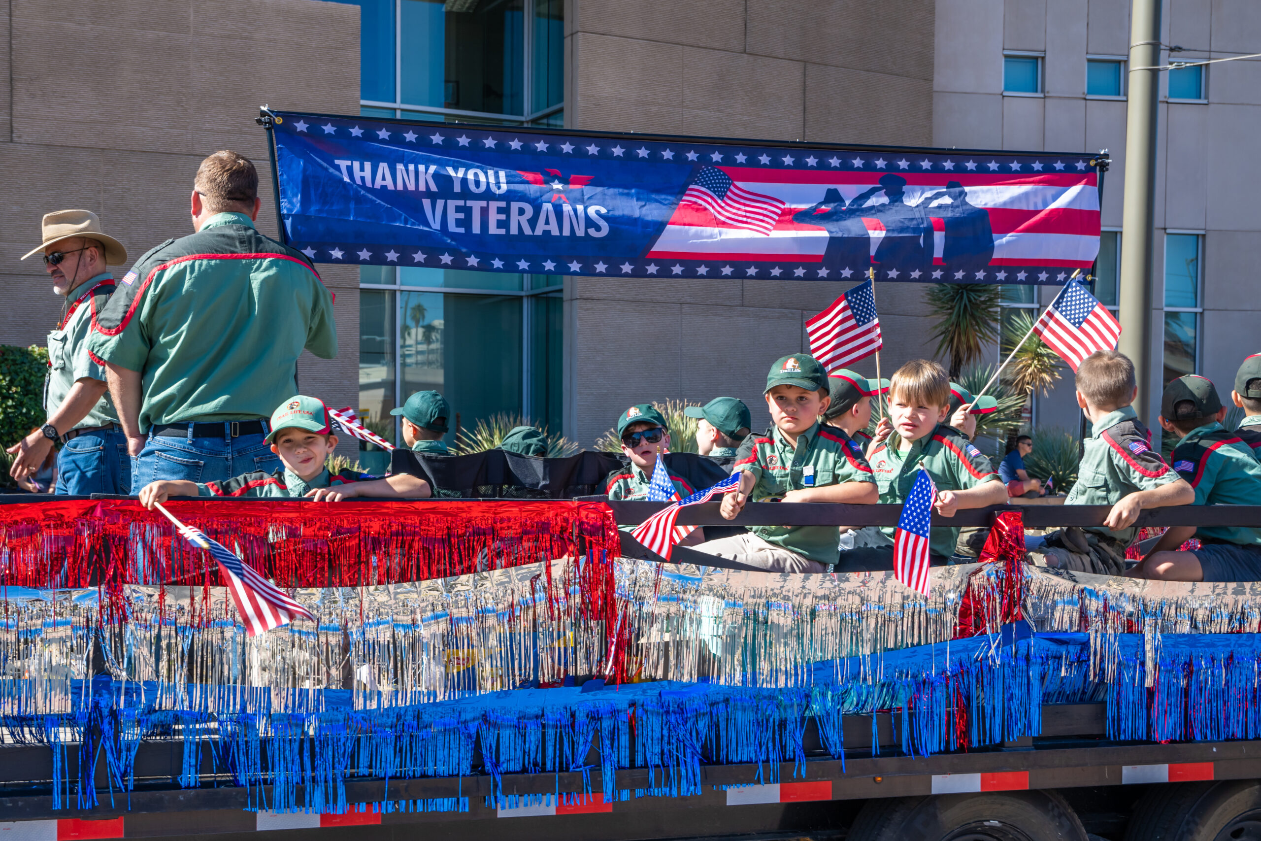 Cub Scouts Thank You Veterans Parade Float Young Cub Scouts in green uniforms waving American flags from a decorated parade float with a "Thank You Veterans" banner.