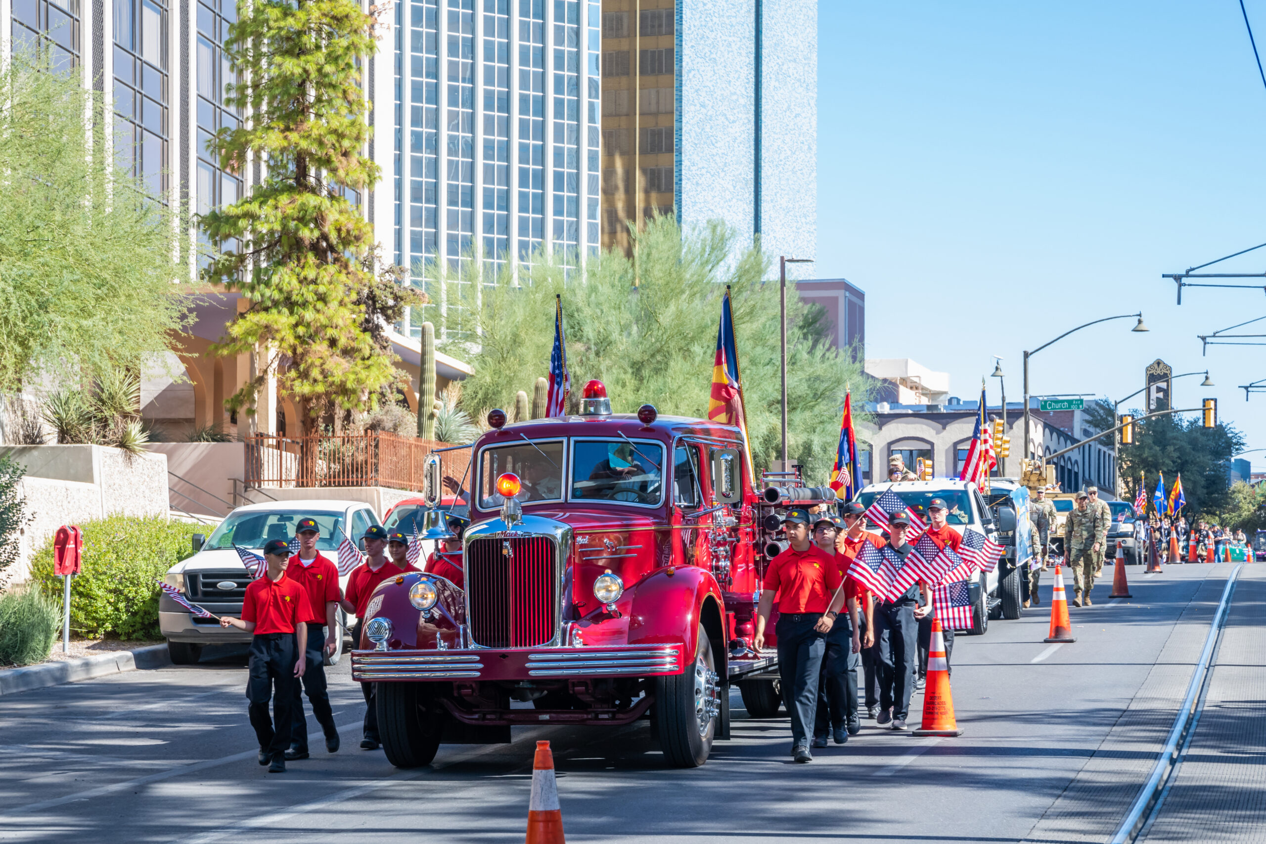 Red Vintage Fire Truck in Downtown Parade A vintage red fire engine leading a parade down a city street, accompanied by young men in red shirts carrying American flags.