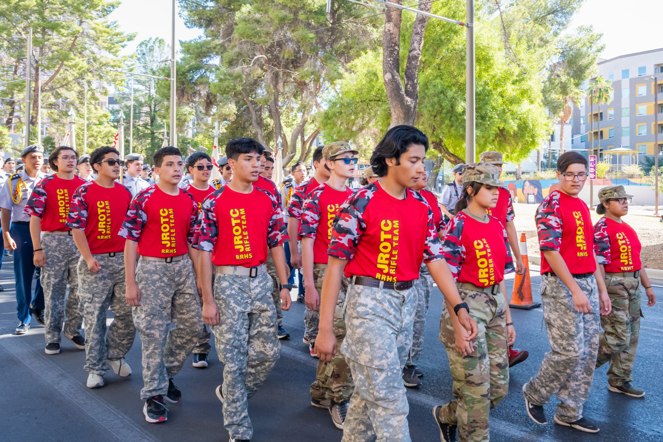 RRHS JROTC Rifle and Raider Team Parade March A formation of students in red shirts with "RRHS JROTC Rifle Team" and "Raider Team" printed on them, marching in a parade wearing camouflage pants.