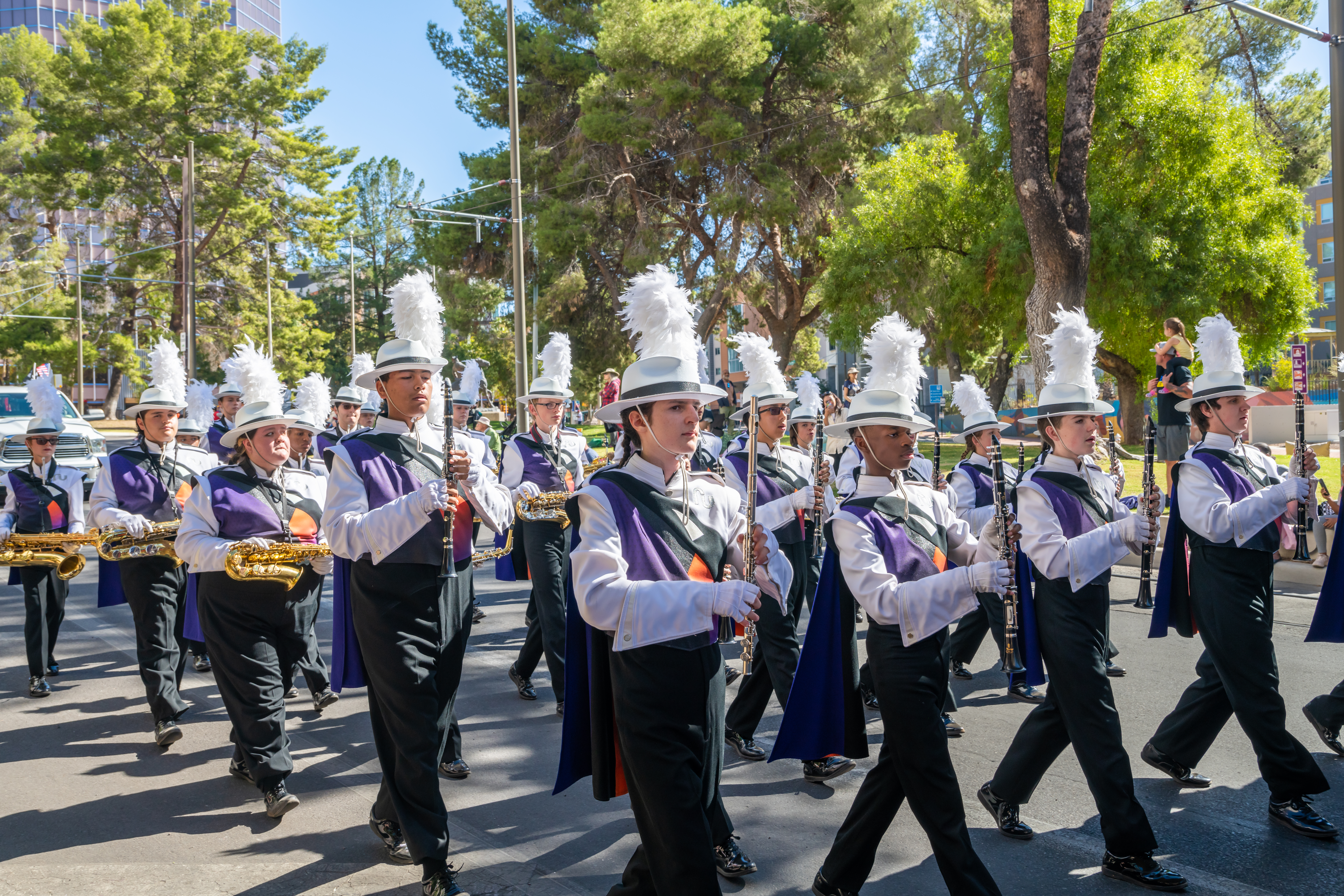 High School Marching Band Performing in Parade A marching band in purple, white, and black uniforms with plumed hats playing saxophones and clarinets in a sunny outdoor parade.