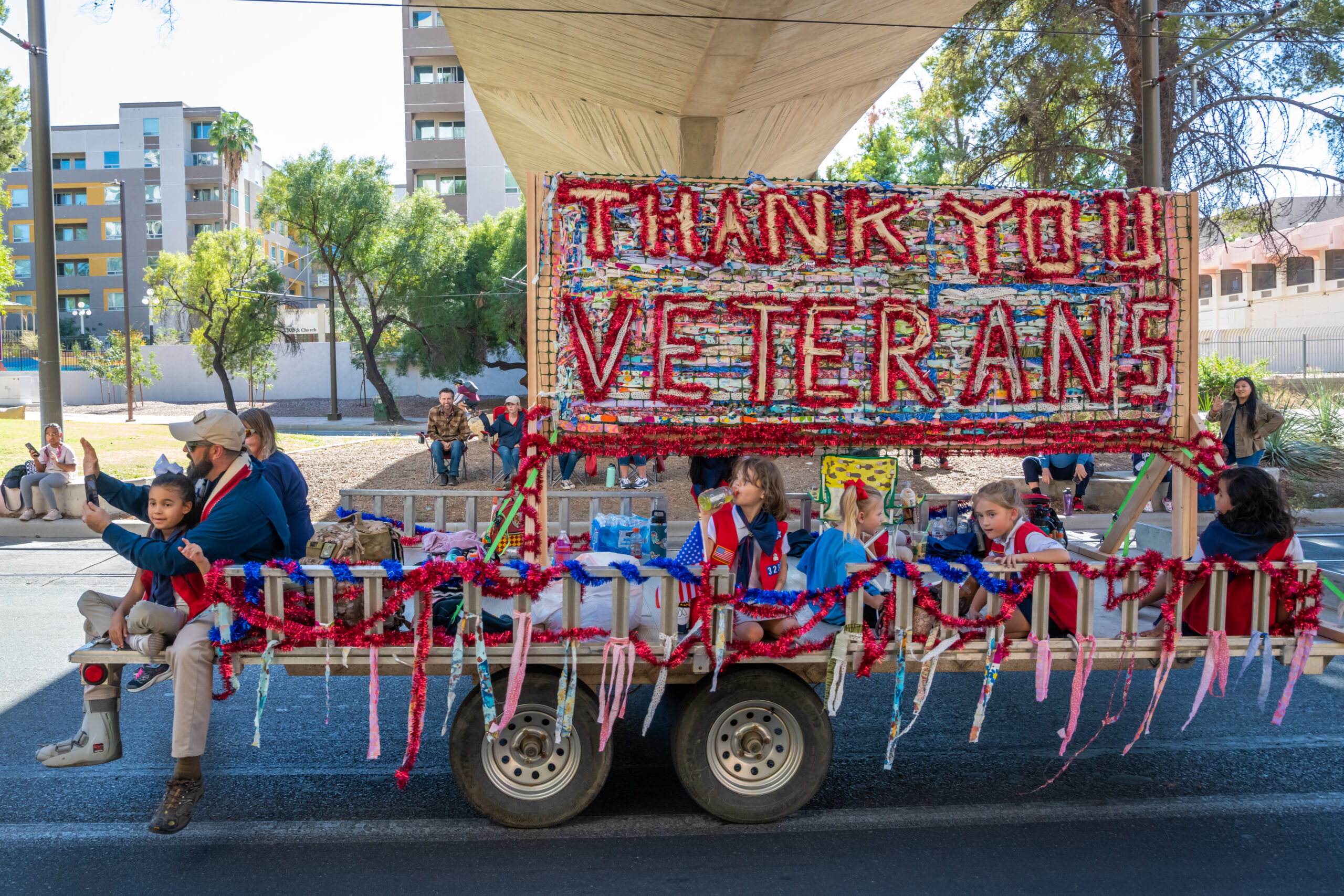 Veterans Day Parade Float with Thank You Sign A parade float decorated with red, white, and blue tinsel and a large handmade "Thank You Veterans" sign, carrying children and a veteran in a walking boot.