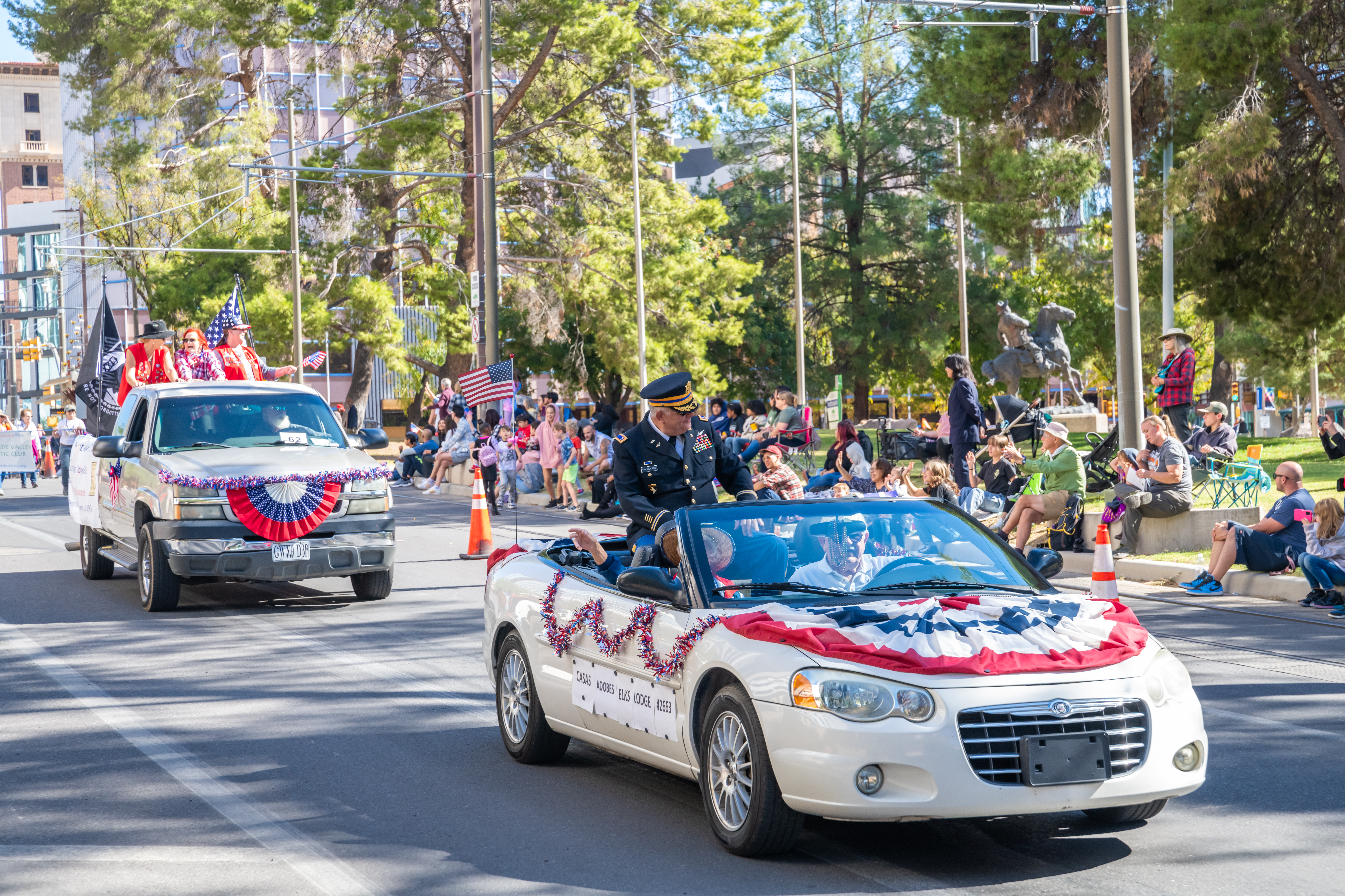 Military Officer Waving from Convertible at Veterans Day Parade A high-ranking military officer in a dress uniform waves to a crowd from a white convertible decorated with American flag bunting during a sunny city parade.