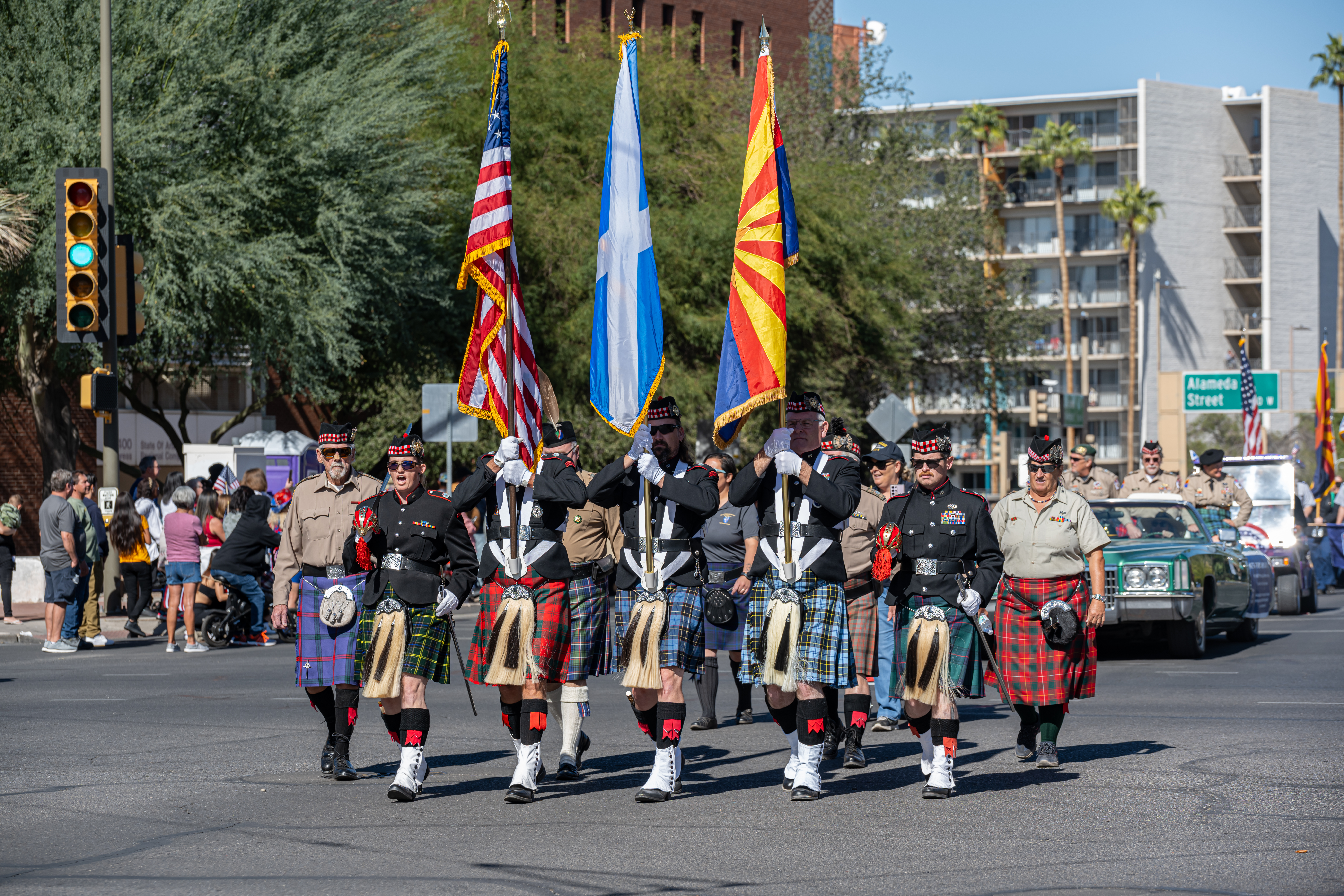 Kilted color guard marching parade A kilted color guard marching down a city street during a parade. They are wearing traditional Scottish tartans, black jackets, and white spats while carrying the American flag, the Arizona state flag, and a blue and white flag.