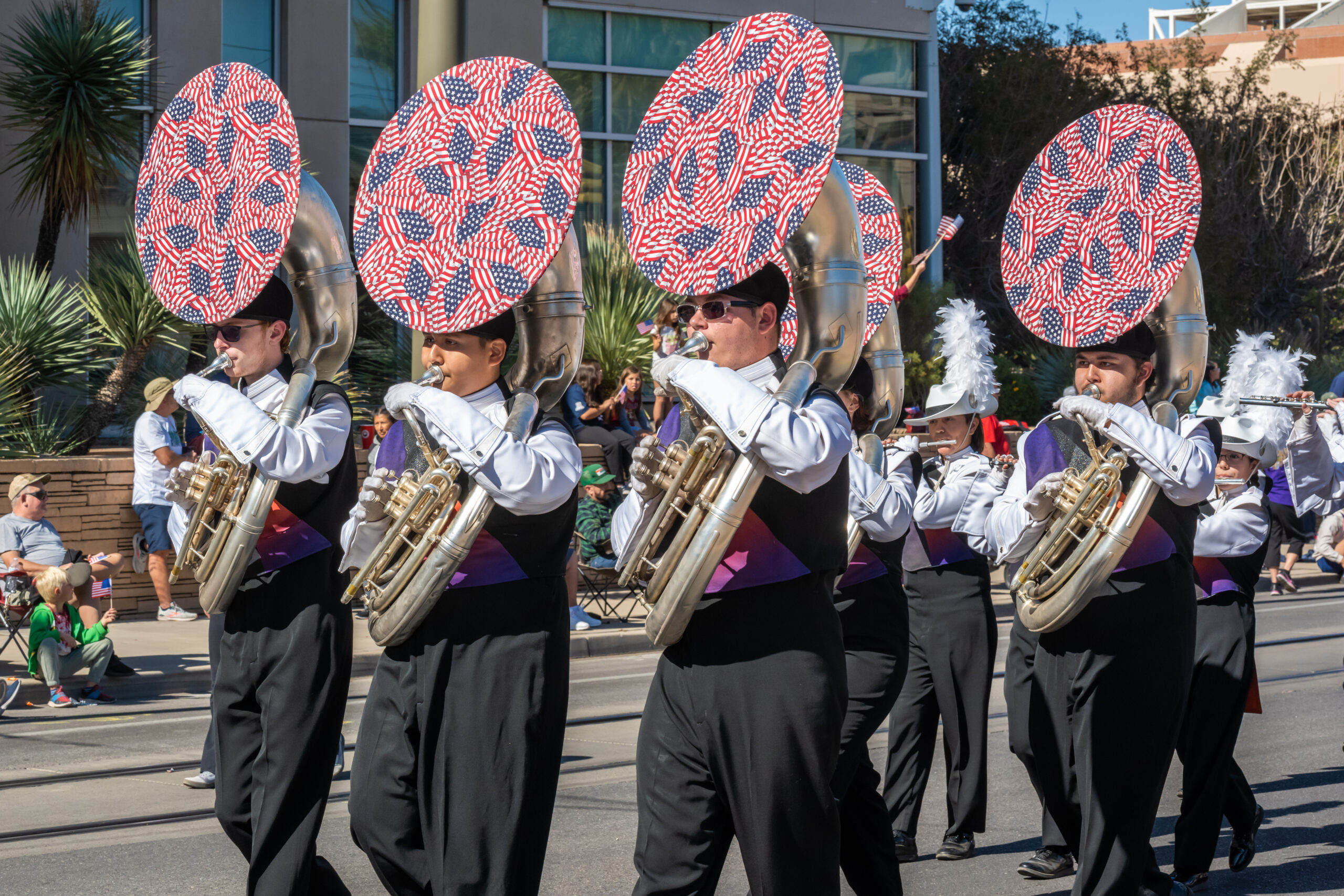Marching band sousaphone section patriotic covers Four marching band members playing silver sousaphones during an outdoor parade. The instruments feature decorative bell covers with a red, white, and blue American flag pattern.