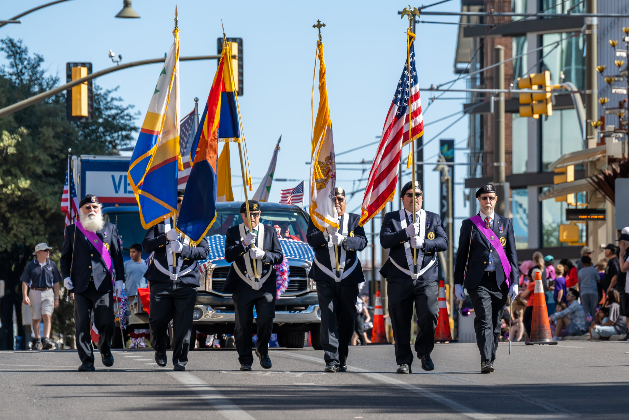 Veterans color guard marching parade tucson A veteran color guard in navy blue blazers and purple sashes marching down a city street in a parade. They are carrying the American flag, the Arizona state flag, and other ceremonial flags while leading a procession.