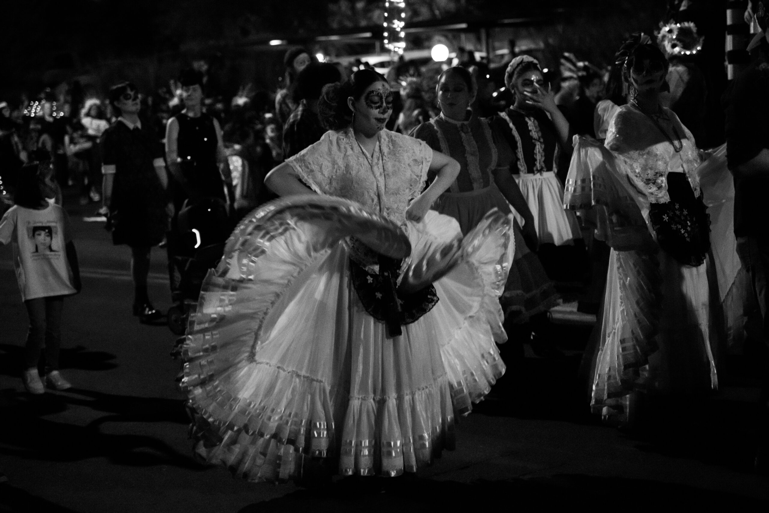 Dia de los Muertos Folklorico Dance A woman with traditional sugar skull face paint performs a folklorico dance, lifting her wide, ruffled white skirt, during a nighttime parade or festival in black and white.