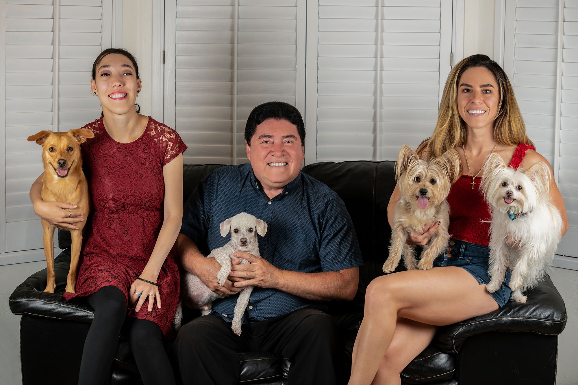Three smiling people sit together on a black leather couch, each holding or sitting with one of four small, happy dogs ranging from a tan pup to a fluffy white one.